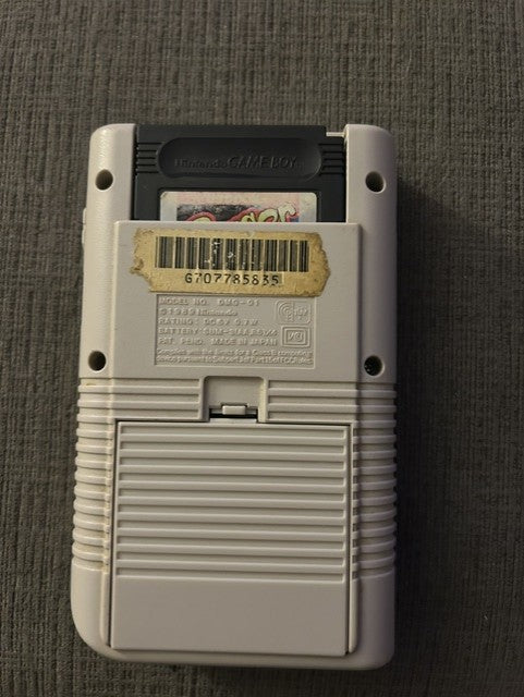 Rear of classic gray Nintendo Game Boy Unit D with worn barcode sticker and battery cover